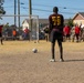 A soccer player wearing jersey number 79 with 'BA' lettering stands facing a goal, preparing to take a penalty kick on a dirt field. A goalkeeper is positioned in the goal with arms outstretched, while a crowd of spectators — many appearing to be in military uniforms — lines the sideline. The setting appears to be a recreational or intramural soccer match at a military installation.
