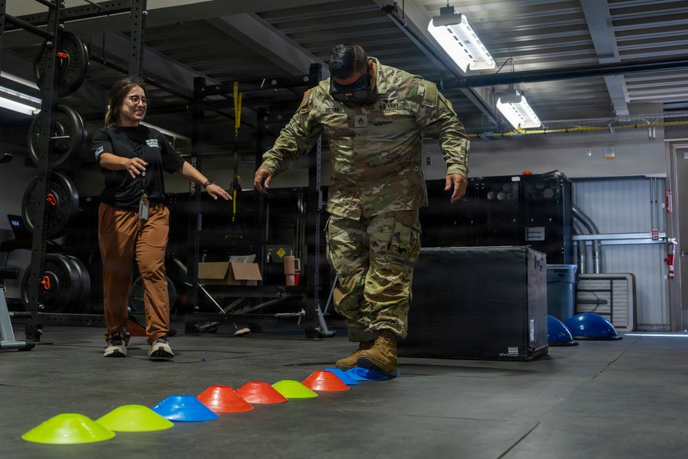 A U.S. Army soldier in full OCP (Operational Camouflage Pattern) uniform is performing an agility drill by stepping through a line of colored marker cones on a gym floor. A civilian instructor or trainer in a black shirt and brown pants is observing and guiding the soldier with an encouraging demeanor. The setting appears to be a military fitness or rehabilitation facility equipped with weight racks and training equipment.