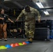 A U.S. Army soldier in full OCP (Operational Camouflage Pattern) uniform is performing an agility drill by stepping through a line of colored marker cones on a gym floor. A civilian instructor or trainer in a black shirt and brown pants is observing and guiding the soldier with an encouraging demeanor. The setting appears to be a military fitness or rehabilitation facility equipped with weight racks and training equipment.