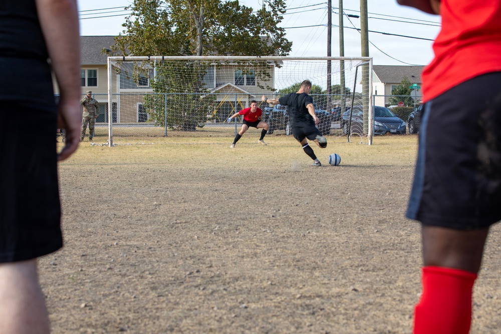 A soccer match is being played on a dry, dirt field near what appears to be a military or residential installation. A player in black is about to kick the ball toward the goal, while a goalkeeper in red prepares to defend. Spectators and bystanders, including a person in military-style camouflage uniform, are visible in the background.