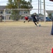 A soccer match is being played on a dry, dirt field near what appears to be a military or residential installation. A player in black is about to kick the ball toward the goal, while a goalkeeper in red prepares to defend. Spectators and bystanders, including a person in military-style camouflage uniform, are visible in the background.