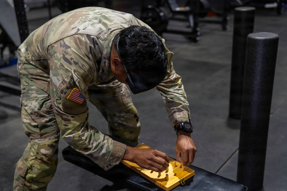 A U.S. Army soldier in OCP (Operational Camouflage Pattern) uniform is leaning over a gym bench, manipulating what appears to be a wooden pegboard or puzzle device. The soldier is wearing a smartwatch and has a face mask pulled down around their neck. The setting is an indoor military fitness facility with gym equipment visible in the background.