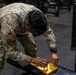 A U.S. Army soldier in OCP (Operational Camouflage Pattern) uniform is leaning over a gym bench, manipulating what appears to be a wooden pegboard or puzzle device. The soldier is wearing a smartwatch and has a face mask pulled down around their neck. The setting is an indoor military fitness facility with gym equipment visible in the background.