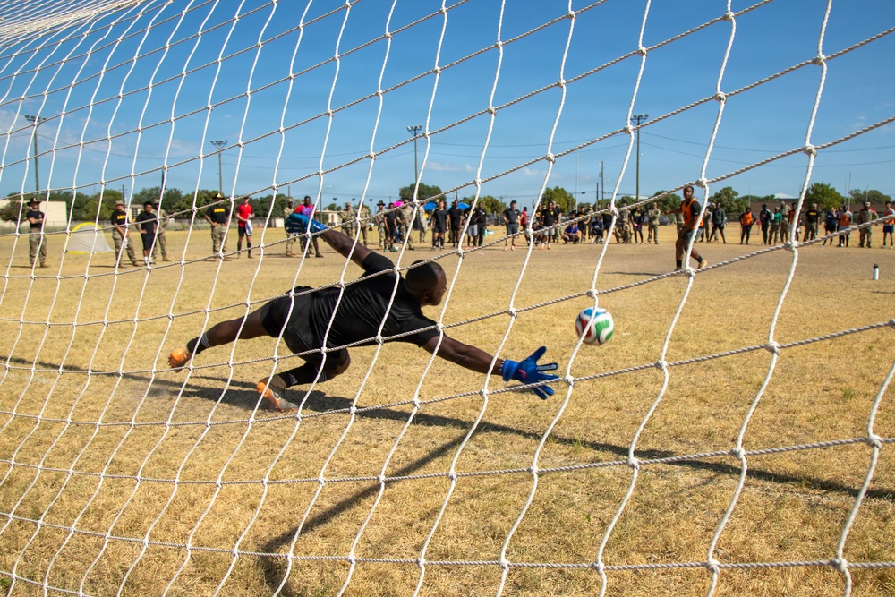 A soccer goalkeeper dressed in black is diving to his right attempting to save a penalty kick, photographed from behind the goal net. The image is taken from inside the goal, giving a unique perspective through the white netting. A crowd of spectators lines the background of a dry grass field under a clear blue sky.