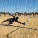 A soccer goalkeeper dressed in black is diving to his right attempting to save a penalty kick, photographed from behind the goal net. The image is taken from inside the goal, giving a unique perspective through the white netting. A crowd of spectators lines the background of a dry grass field under a clear blue sky.