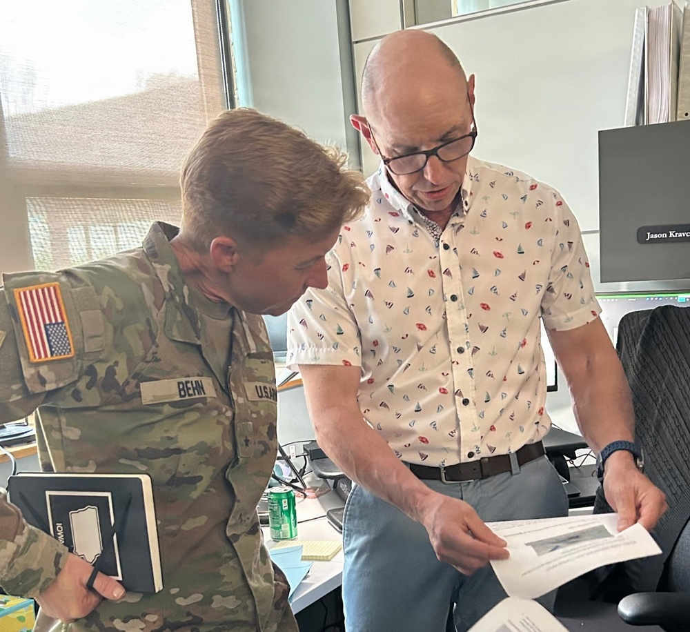 A U.S. Army soldier wearing OCP (Operational Camouflage Pattern) uniform with the name tape 'BEHN' and a U.S. flag patch is reviewing a document with a civilian man in a patterned shirt. They appear to be in an office environment, leaning over a printed document together. The civilian's computer monitor in the background displays the name 'Jason Kravc' (partially visible).