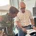 A U.S. Army soldier wearing OCP (Operational Camouflage Pattern) uniform with the name tape 'BEHN' and a U.S. flag patch is reviewing a document with a civilian man in a patterned shirt. They appear to be in an office environment, leaning over a printed document together. The civilian's computer monitor in the background displays the name 'Jason Kravc' (partially visible).