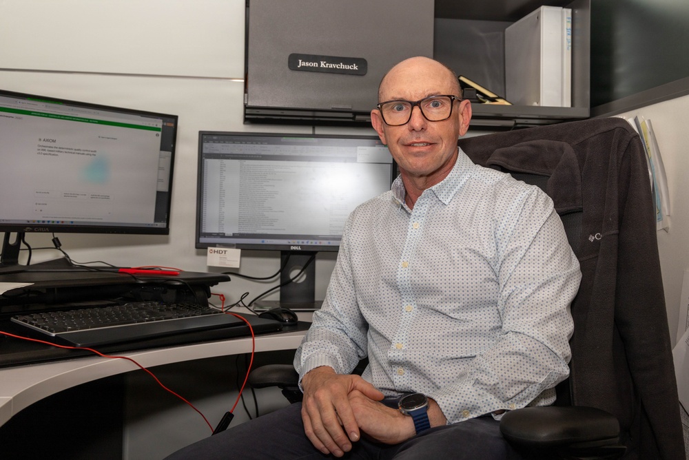 A man identified by a nameplate as Jason Kravchuck is seated at an office workstation, posing for what appears to be a professional or workplace portrait. He is surrounded by dual computer monitors, a keyboard, and office equipment in a cubicle-style workspace. He is dressed in a light blue patterned button-up shirt and wears glasses and a watch.