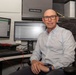 A man identified by a nameplate as Jason Kravchuck is seated at an office workstation, posing for what appears to be a professional or workplace portrait. He is surrounded by dual computer monitors, a keyboard, and office equipment in a cubicle-style workspace. He is dressed in a light blue patterned button-up shirt and wears glasses and a watch.