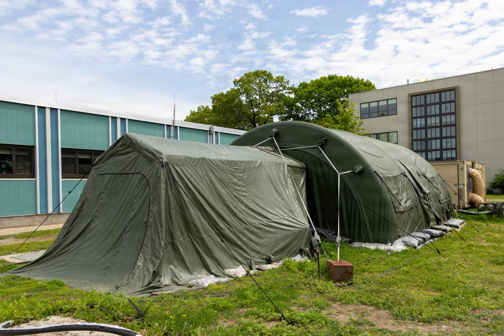 The image shows large military-style inflatable/modular olive-green tents set up on a grass area between institutional buildings. The tents appear to be field hospital or command post structures, anchored with sandbags and guy-wires. The setting suggests a temporary operational deployment adjacent to permanent structures.
