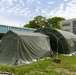 The image shows large military-style inflatable/modular olive-green tents set up on a grass area between institutional buildings. The tents appear to be field hospital or command post structures, anchored with sandbags and guy-wires. The setting suggests a temporary operational deployment adjacent to permanent structures.