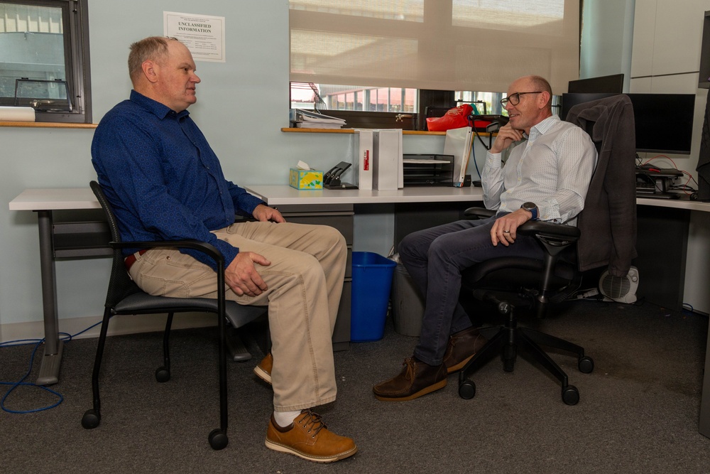 Two men are seated facing each other in an office environment, engaged in what appears to be a professional conversation or meeting. One man wears a blue shirt with khaki pants, while the other wears a light-colored checkered shirt with dark jeans and glasses. A sign reading 'UNCLASSIFIED INFORMATION' is visible on the wall in the background.