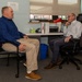Two men are seated facing each other in an office environment, engaged in what appears to be a professional conversation or meeting. One man wears a blue shirt with khaki pants, while the other wears a light-colored checkered shirt with dark jeans and glasses. A sign reading 'UNCLASSIFIED INFORMATION' is visible on the wall in the background.