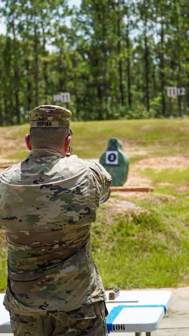 A U.S. Army soldier named Ospina is seen from behind at an outdoor shooting range, aiming a pistol at a close-range silhouette target. The soldier is wearing OCP (Operational Camouflage Pattern) uniform and patrol cap. Additional numbered target stands are visible in the background at greater distances.