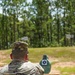 A U.S. Army soldier named Ospina is seen from behind at an outdoor shooting range, aiming a pistol at a close-range silhouette target. The soldier is wearing OCP (Operational Camouflage Pattern) uniform and patrol cap. Additional numbered target stands are visible in the background at greater distances.