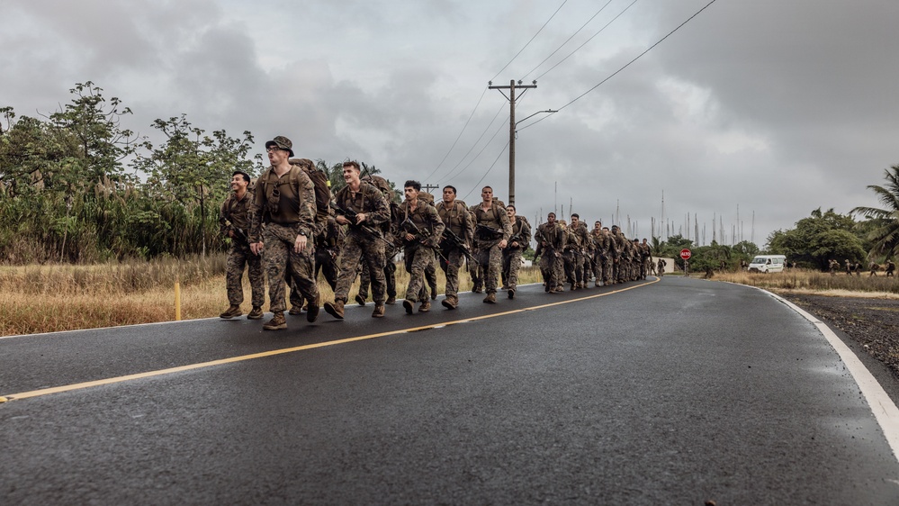 A column of military personnel in camouflage uniforms conducts a road march along a wet paved road in what appears to be a tropical environment. The soldiers are carrying full combat gear including backpacks and rifles. A stop sign reading 'ALTO' is visible in the background, suggesting a Spanish-speaking country or territory.