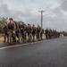 A column of military personnel in camouflage uniforms conducts a road march along a wet paved road in what appears to be a tropical environment. The soldiers are carrying full combat gear including backpacks and rifles. A stop sign reading 'ALTO' is visible in the background, suggesting a Spanish-speaking country or territory.