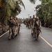 A column of armed military personnel in camouflage uniforms marches along a paved two-lane road flanked by tropical vegetation including palm trees. The soldiers are carrying rifles and wearing tactical gear, moving in formation toward the camera. The setting appears to be a tropical or subtropical region based on the lush greenery and road infrastructure visible.