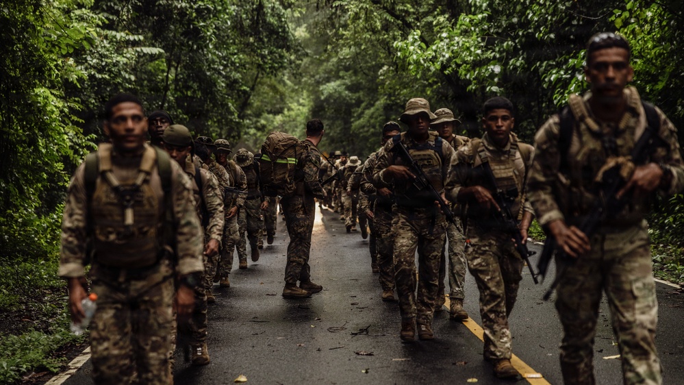 A column of military personnel in camouflage uniforms marches along a wet paved road through dense tropical jungle. The soldiers are equipped with tactical gear, backpacks, and firearms, suggesting a training exercise or patrol operation. The lush green canopy overhead and wet road surface indicate a humid, rainy tropical environment.