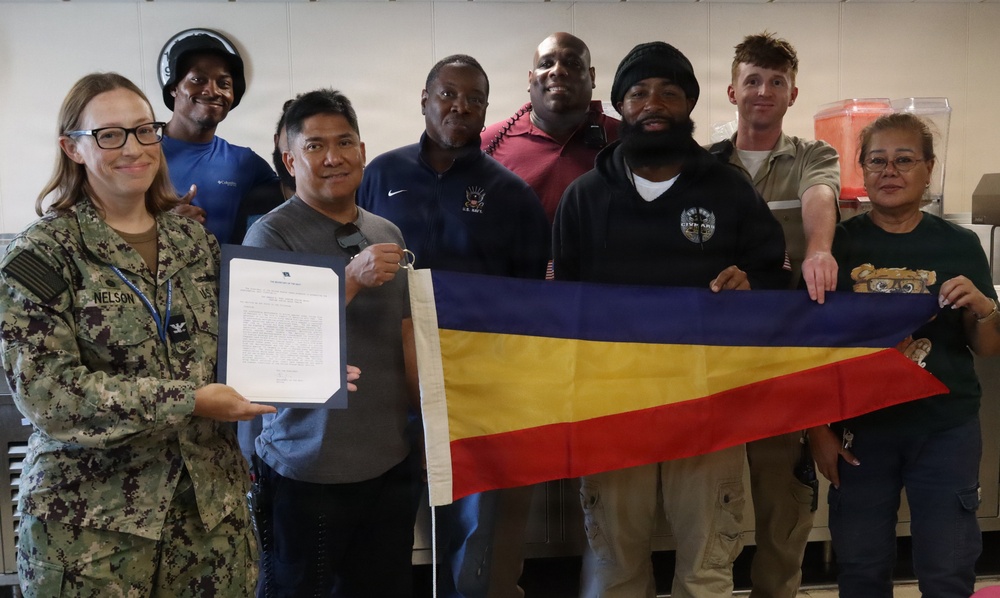 A group of approximately eight individuals pose for a formal photo, with one person in U.S. Navy camouflage uniform (name tape reads 'NELSON') holding an official document. Several members of the group are holding a pennant-style flag with horizontal blue, yellow, and red stripes. The setting appears to be an indoor facility, likely a military or government workplace.