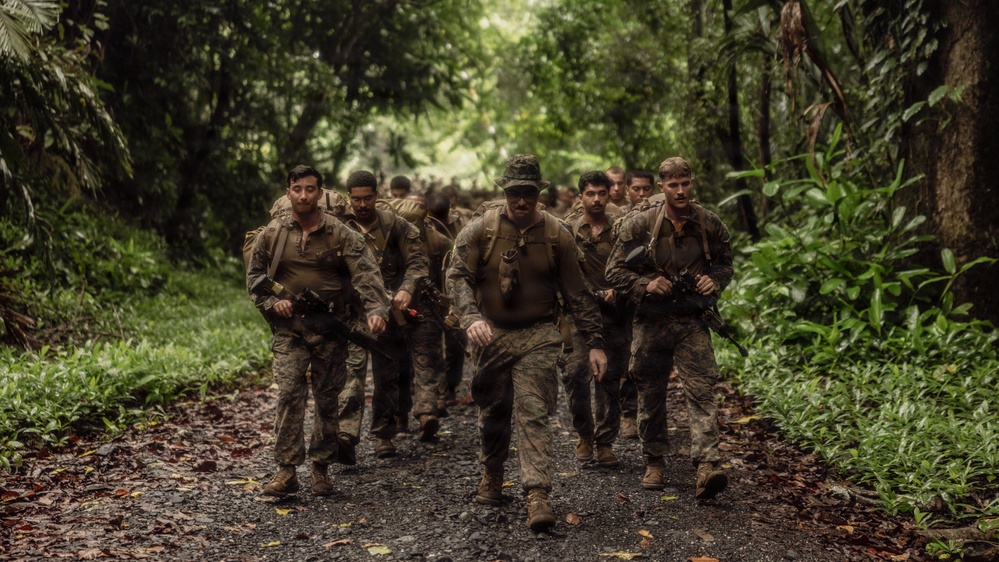 A group of military personnel in camouflage uniforms march in formation along a narrow jungle path. The soldiers appear fatigued and mud-covered, suggesting they have been conducting extended field operations in a tropical environment. The column extends deep into the background, indicating a sizeable unit on patrol or a training exercise.