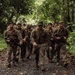 A group of military personnel in camouflage uniforms march in formation along a narrow jungle path. The soldiers appear fatigued and mud-covered, suggesting they have been conducting extended field operations in a tropical environment. The column extends deep into the background, indicating a sizeable unit on patrol or a training exercise.