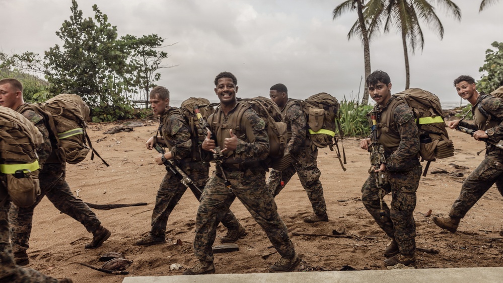 A group of military personnel in camouflage uniforms and full combat gear are marching across a sandy beach area with tropical vegetation in the background. The soldiers are carrying large rucksacks and rifles, with several appearing in good spirits, smiling and giving thumbs up. The setting appears to be a tropical coastal environment, consistent with a training exercise or field operation.