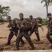 A group of military personnel in camouflage uniforms and full combat gear are marching across a sandy beach area with tropical vegetation in the background. The soldiers are carrying large rucksacks and rifles, with several appearing in good spirits, smiling and giving thumbs up. The setting appears to be a tropical coastal environment, consistent with a training exercise or field operation.