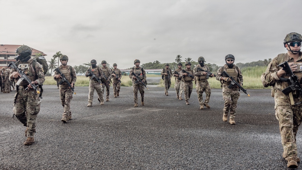 A group of approximately 10-15 armed military or special operations personnel in full tactical gear are walking across what appears to be a tarmac or airfield surface. All individuals are equipped with assault rifles, body armor, helmets, and camouflage uniforms. The setting appears to be a tropical location based on the palm trees and vegetation visible in the background.