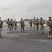 A group of approximately 10-15 armed military or special operations personnel in full tactical gear are walking across what appears to be a tarmac or airfield surface. All individuals are equipped with assault rifles, body armor, helmets, and camouflage uniforms. The setting appears to be a tropical location based on the palm trees and vegetation visible in the background.