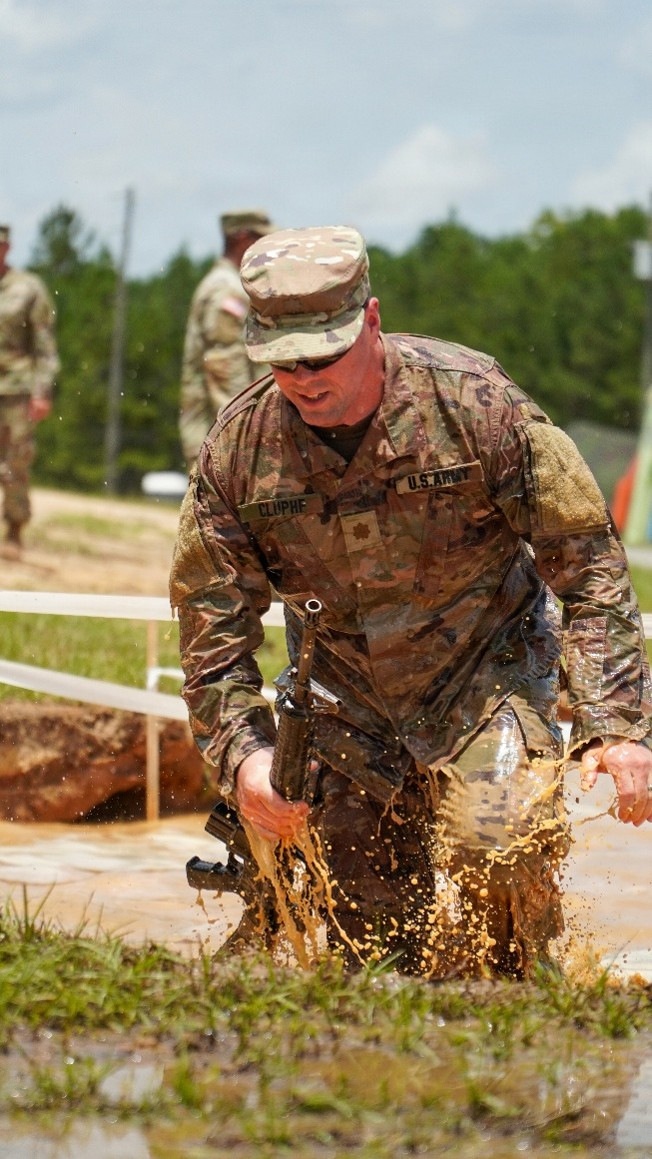 A U.S. Army soldier wearing OCP (Operational Camouflage Pattern) uniform with the name tape 'CLUPHF' and a rank insignia is navigating through a muddy water obstacle, likely during a military training exercise or competition. The soldier is soaking wet and splashing through muddy water while carrying what appears to be a rifle. Additional soldiers in uniform are visible in the background observing or participating.