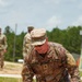 A U.S. Army soldier wearing OCP (Operational Camouflage Pattern) uniform with the name tape 'CLUPHF' and a rank insignia is navigating through a muddy water obstacle, likely during a military training exercise or competition. The soldier is soaking wet and splashing through muddy water while carrying what appears to be a rifle. Additional soldiers in uniform are visible in the background observing or participating.