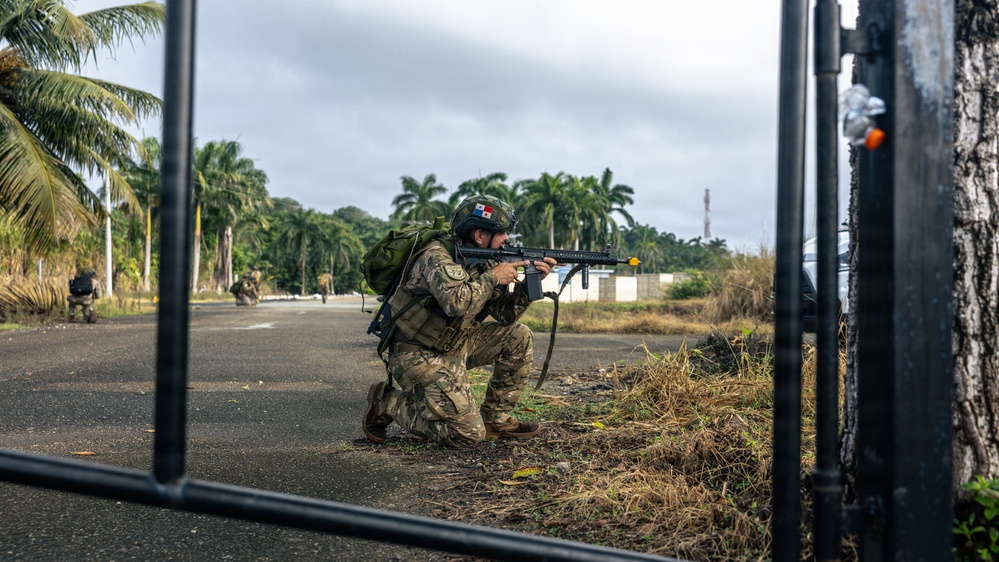 A soldier in full combat gear is kneeling in a tactical firing position near a gate, aiming a rifle down a road flanked by palm trees. A flag patch visible on the helmet appears to be the Panamanian flag, suggesting this may be a Panamanian military or security forces exercise. Additional personnel in tactical gear are visible in the background along the road.