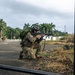 A soldier in full combat gear is kneeling in a tactical firing position near a gate, aiming a rifle down a road flanked by palm trees. A flag patch visible on the helmet appears to be the Panamanian flag, suggesting this may be a Panamanian military or security forces exercise. Additional personnel in tactical gear are visible in the background along the road.
