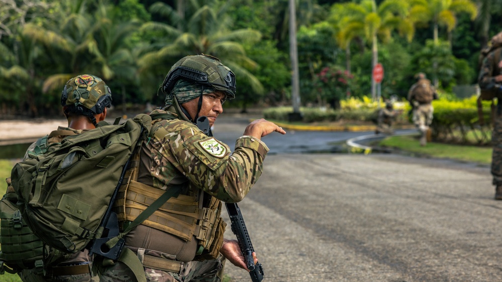 The image shows military or special operations personnel in a tropical environment, likely during a training exercise or patrol operation. A central figure in multicam camouflage with a tactical vest, helmet, and rifle is gesturing/pointing toward a direction, appearing to give instructions. Additional personnel are visible in the background along a paved road lined with palm trees and tropical vegetation.