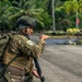 The image shows military or special operations personnel in a tropical environment, likely during a training exercise or patrol operation. A central figure in multicam camouflage with a tactical vest, helmet, and rifle is gesturing/pointing toward a direction, appearing to give instructions. Additional personnel are visible in the background along a paved road lined with palm trees and tropical vegetation.