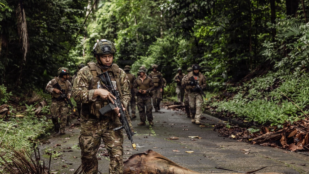 A group of armed military personnel in tactical gear are conducting a patrol along a narrow paved path through a dense tropical jungle environment. The lead soldier is wearing multicam camouflage and carrying an assault rifle, with additional personnel following in a loose column formation. At least one soldier in the background appears to display a Panamanian flag patch on their uniform.