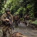 A group of armed military personnel in tactical gear are conducting a patrol along a narrow paved path through a dense tropical jungle environment. The lead soldier is wearing multicam camouflage and carrying an assault rifle, with additional personnel following in a loose column formation. At least one soldier in the background appears to display a Panamanian flag patch on their uniform.
