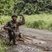 A soldier in full combat gear is positioned at the edge of a paved path bordered by dense tropical vegetation. The soldier is crouching near tree cover and appears to be giving a hand signal, likely communicating with other personnel. A second figure in military uniform is faintly visible in the background, suggesting a small unit tactical exercise or patrol.