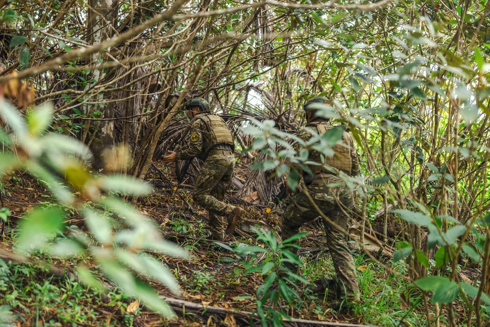 Two individuals in military-style camouflage uniforms and tactical gear are moving through dense jungle or forest vegetation. Both are wearing helmets and plate carriers/vests with visible patches, navigating through thick brush and undergrowth. The image appears to capture a patrol or training exercise in a tropical or subtropical environment.