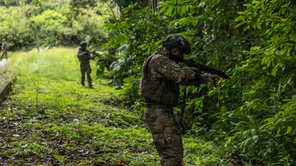 Two soldiers in multicam camouflage uniforms are conducting a patrol or training exercise along a jungle trail. The foreground soldier is in an aiming stance with a rifle, while a second soldier is visible further down the path. A small flag patch is visible on the foreground soldier's helmet, identifiable as the Panamanian flag.