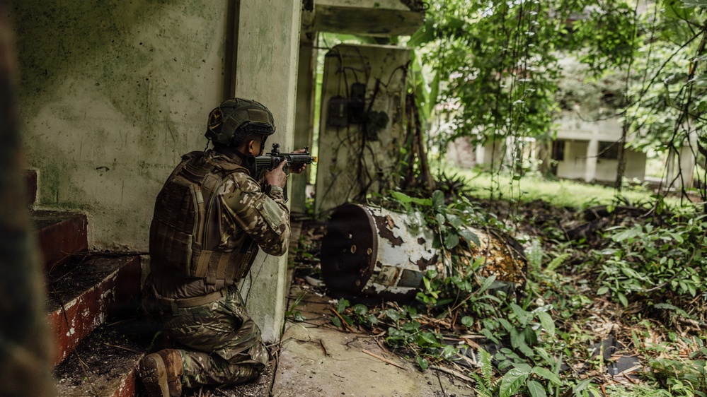 A soldier in full camouflage gear and tactical helmet is kneeling in a firing position at the corner of a deteriorating building, aiming a rifle outward. The environment appears to be an overgrown, abandoned compound with dense tropical vegetation. The scene suggests a military training exercise or operational patrol in an urban-jungle environment.