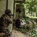 A soldier in full camouflage gear and tactical helmet is kneeling in a firing position at the corner of a deteriorating building, aiming a rifle outward. The environment appears to be an overgrown, abandoned compound with dense tropical vegetation. The scene suggests a military training exercise or operational patrol in an urban-jungle environment.