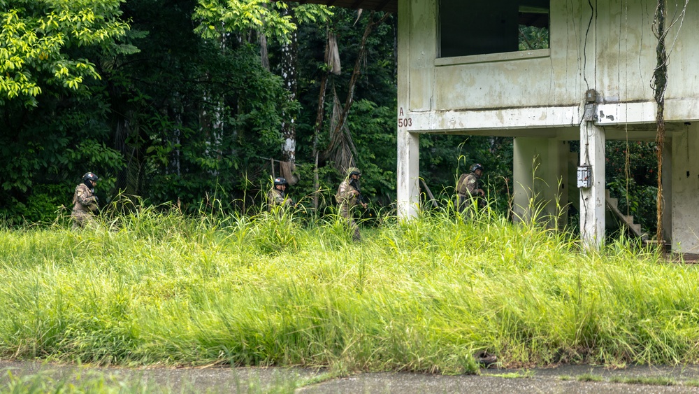 The image shows four individuals in military or tactical gear moving through tall grass near an abandoned or dilapidated two-story concrete building marked 'A 503'. The personnel are wearing helmets and camouflage uniforms and appear to be conducting a tactical training exercise or patrol in an overgrown area. The setting suggests a military training facility with deteriorating infrastructure surrounded by dense tropical or subtropical vegetation.