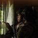 A soldier in full tactical gear is positioned inside a building, looking out through a window while pointing toward an exterior location. The individual is equipped with a combat helmet fitted with accessories, camouflage uniform, communications headset, and is carrying a firearm. The scene appears to depict a military reconnaissance or urban operations scenario.
