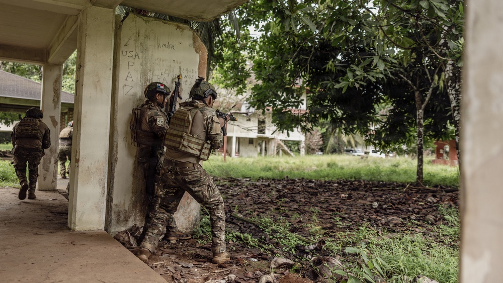 The image shows armed military or law enforcement personnel conducting what appears to be a tactical clearing operation around a deteriorated concrete structure. Two soldiers in the foreground are stacked against a corner wall in a tactical position, while at least one additional personnel is visible in the background moving along a covered walkway. The environment appears to be a partially abandoned or damaged compound with lush tropical vegetation.