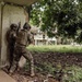 The image shows armed military or law enforcement personnel conducting what appears to be a tactical clearing operation around a deteriorated concrete structure. Two soldiers in the foreground are stacked against a corner wall in a tactical position, while at least one additional personnel is visible in the background moving along a covered walkway. The environment appears to be a partially abandoned or damaged compound with lush tropical vegetation.