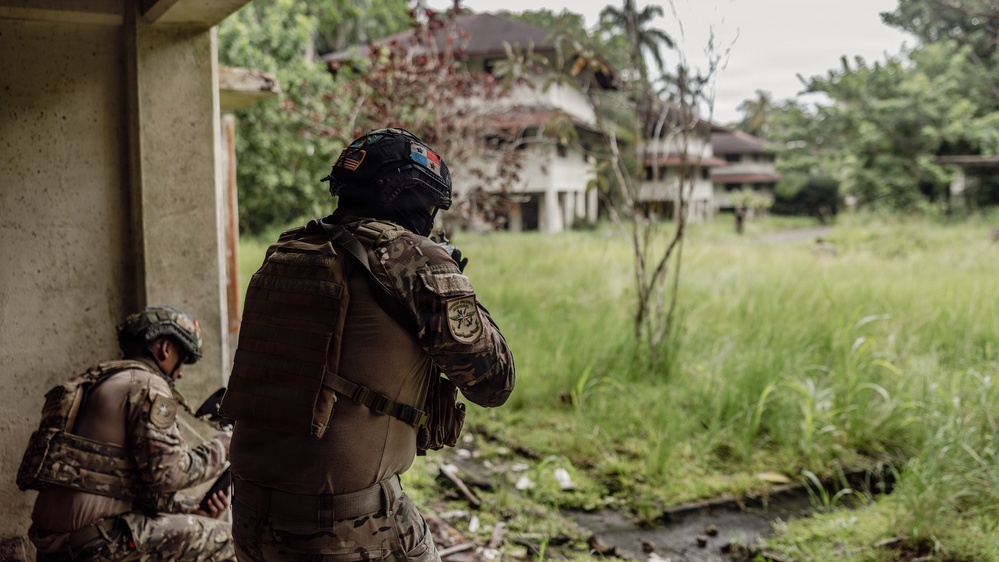 Two armed military or law enforcement personnel in full tactical gear are positioned near a concrete structure, observing an abandoned or dilapidated building across an overgrown field. The foreground operator wears a black helmet with what appears to be flag patches and a tactical vest with unit insignia. The setting appears to be a tropical environment based on the lush vegetation and palm trees visible in the background.
