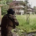 Two armed military or law enforcement personnel in full tactical gear are positioned near a concrete structure, observing an abandoned or dilapidated building across an overgrown field. The foreground operator wears a black helmet with what appears to be flag patches and a tactical vest with unit insignia. The setting appears to be a tropical environment based on the lush vegetation and palm trees visible in the background.