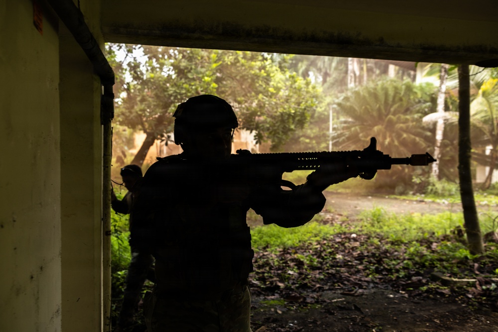 A silhouetted figure in tactical gear and helmet is aiming a rifle from inside a structure toward a lush, tropical outdoor environment. A second figure is partially visible behind them near a wall. The scene appears to depict a military or law enforcement training exercise in a tropical setting.